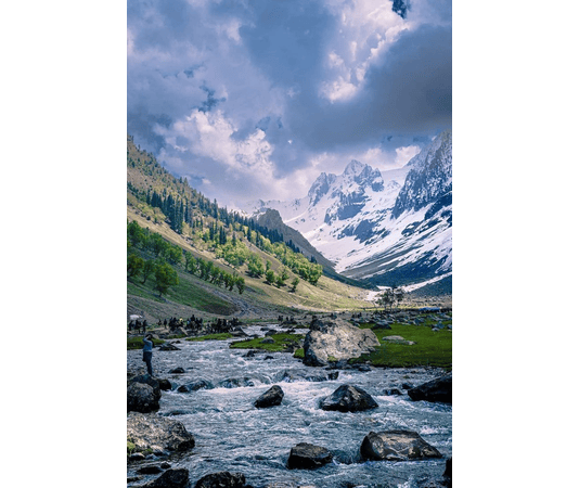 A breathtaking view of a crystal-clear river flowing through a lush green valley, surrounded by towering snow-capped mountains under a dramatic sky.